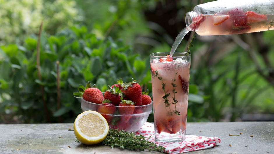 Pouring a soda into glass, bowl of strawberries and a lemon slice in background 